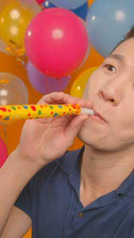 Vertical-Video-Studio-Portrait-Of-Man-Wearing-Party-Hat-Celebrating-Birthday-With-Balloons-And-Party-Blower-1